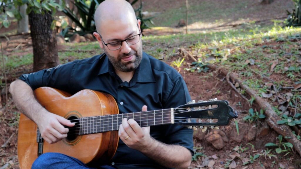 3a foto de Guilherme Mauad, membro do Duo Origens, tocando violão 6 cordas sentado em uma praça
